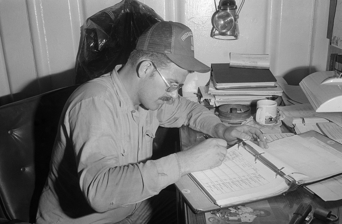 A man, middle-aged, White, wearing a baseball-style cap and weathered work shirt, sitting at a desk full of papers and writing in a 3-ring binder.